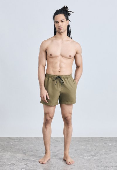 Άνδρας με μακριά dreadlocks στέκεται ξυπόλητος μπροστά σε γκρι τοίχο, φορώντας χακί πράσινα μαγιό με μαύρο κορδόνι, με το ένα χέρι στην τσέπη.