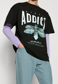 Černé oversized tričko s nápisem "NIGHT ADDICT" a grafikou můry, vrstvené přes fialové dlouhé tričko; světle modré kalhoty.