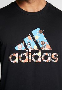 Schwarzes Baumwoll-T-Shirt mit einem bunten pixelierten Adidas-Logo, das verschiedene Sportikonen, darunter Bälle und Ausrüstung in Blau, Orange und Gelb, zeigt.