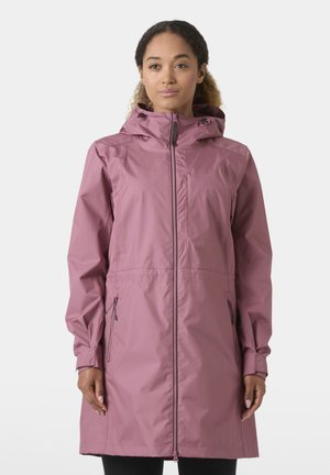 Γυναίκα που φοράει μακριά αδιάβροχη παλτό σε αποχρώσεις μωβ με κουκούλα, φερμουάρ μπροστά και φερμουάρ στις πλαϊνές τσέπες, στέκεται μπροστά σε απλό φόντο.