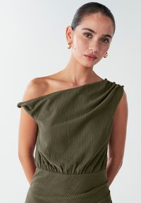 Olijfgroen, gestructureerd een-schouder top met geplooide details bij de schouder en taille. Gouden oorbellen accentueren de outfit.