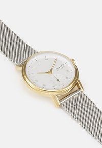 Zlatý hodinky s stříbrným pružinovým páskem. Mají bílý ciferník s černými indexy, zlatými ručičkami a malou subciferníkem na šesti hodin.