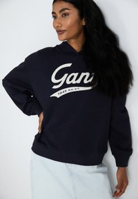 Navy blue hoodie with a front kangaroo pocket and 'Gant' graphic in off-white. Constructed from soft fabric with a relaxed fit.
