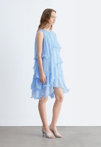 Femme portant une robe bleu clair sans manches, composée de plusieurs couches, et des sandales argentées à talons, debout sur un sol gris devant un mur blanc.
