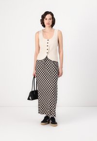 Gilet in lino beige con chiusura a bottoni, abbinato a una gonna maxi a quadri bianca e nera. La modella tiene una piccola borsa nera e indossa sneakers nere.