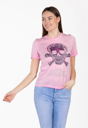 Kobieta w różowej koszulce z cekinowym wzorem czaszki i piszczeli oraz tekstem, w połączeniu z jasnoniebieskimi dżinsami, uśmiechająca się i pozująca z jedną ręką w kieszeni.