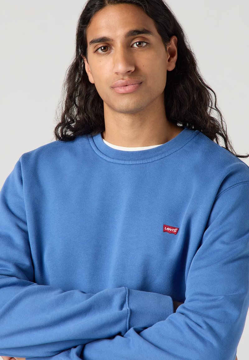 Sweatshirt bleu avec un col rond, des manches longues et un petit logo rouge Levi's sur la poitrine. Tissu doux avec une texture lisse.