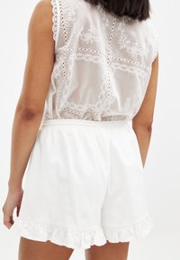 Białe lniane szorty z falbaniastym dołem, połączone z przezroczystą, haftowaną bluzką z kwiatowymi wzorami i detalami w oczkach na ramionach.