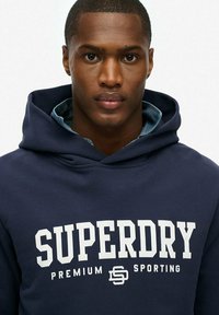 Marineblauwe hoodie met een groot wit "SUPERDRY" logo, voorzien van een capuchon met veters, zachte stof, en een gelaagde denim shirt zichtbaar bij de kraag.