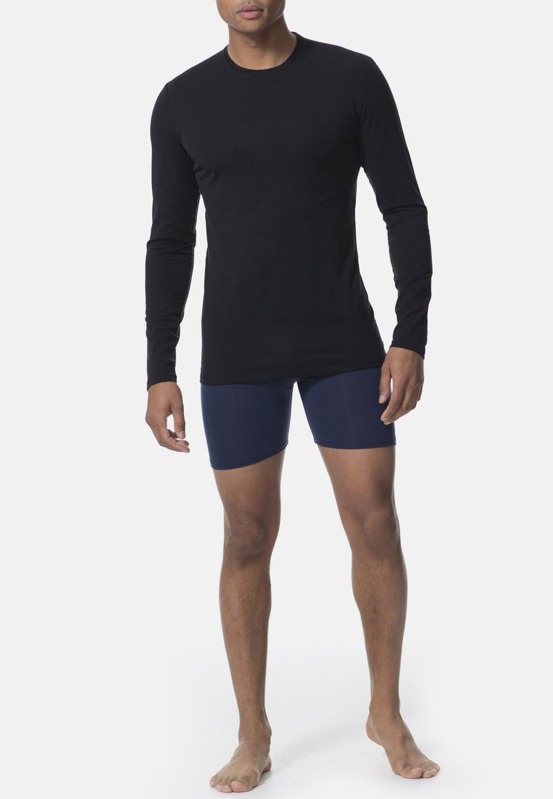 Black long-sleeve shirt made of smooth fabric, paired with navy blue fitted shorts. Model standing barefoot, hands relaxed by sides.
