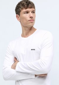 Bijela majica dugih rukava s okruglim izrezom i malim crnim logom "BOSS" na lijevoj strani grudi. Glatka pamučna tekstura.