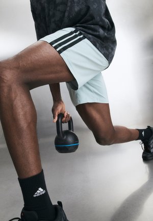 Muškarac u sportskoj opremi radi iskorak držeći crnu kettlebell s plavom prugom na glatkom podu.