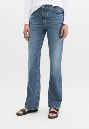 Persoon die blauwe rechte jeans draagt, een witte top met lange mouwen en zwarte open sandalen, staand tegen een effen achtergrond.