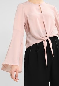 Vaaleanpunainen paita, jossa on pyöreä pääntie, avainreikäyksityiskohta ja pitkät kellohihat; solmittu vyötärölle pehmeällä, teksturoidulla kankaalla. yhdistetty mustiin housuihin.