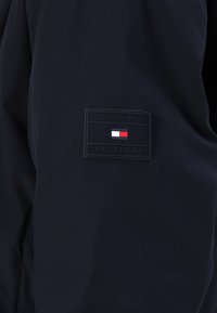 Giacca blu marina con un logo rettangolare nero sulla manica, che presenta il testo "TOMMY HILFIGER" e un piccolo elemento grafico in rosso, bianco e blu.