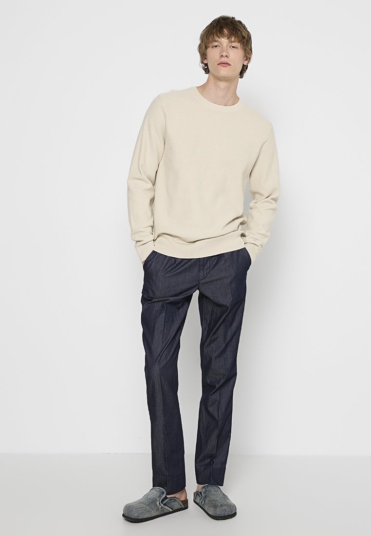 Beige texturerad sweatshirt, mörkblå avsmalnande byxor, grå slip-on skor. Kläderna har en avslappnad och bekväm passform.