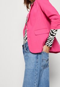 Světle růžový blazer s přizpůsobeným designem, obsahující dva knoflíky a jedno náprsní kapsu, překrytý černobílým topem se zebrovaným vzorem.
