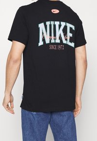 Schwarzes Baumwoll-T-Shirt mit einem großen "NIKE"-Grafik in Türkis und Pink, darunter der Schriftzug "Champions of Sport" und "Seit 1972".