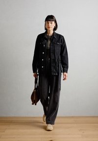Giubbotto di denim nero, top verde oliva e pantaloni neri larghi. Porta una borsa marrone, indossa scarpe beige. Design semplice, texture naturali.