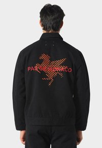 Schwarze Jacke mit einem strukturierten orangen Design eines Pferdes und dem Text "PAS DE MONACO" auf dem Rücken; lange Ärmel und klassischer Kragen.