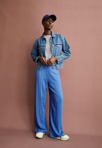 Denim jacket, grey t-shirt, blue wide-leg pants, and white sneakers with green accents. Model poses against a pastel pink background.