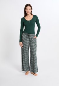 Vrouw die blootvoets staat, gekleed in een donkergroene lange mouwtop en losse geruite broek, met een hand in de zak tegen een witte achtergrond.