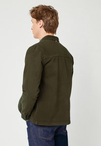 Dark green casual jacket with long sleeves, buttoned cuffs, and back yoke detail. Worn over blue jeans. Smooth texture, simple design.