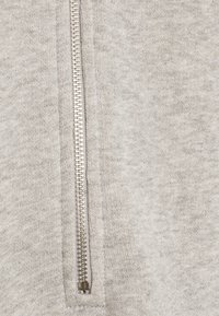 Sudadera de cremallera en gris claro con una cremallera metálica, que muestra una tela suave y texturada y un diseño limpio. La cremallera se encuentra en el centro de la parte delantera.