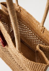 Geflochtener Strohtaschen mit dicken Griffen, bestehend aus einem offenen Geflechtmuster, in hellbrauner Farbe und mit weicher Innenauskleidung. Enthält ein farbenfrohes Quasten-Detail.