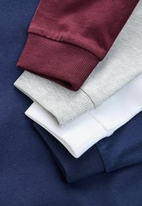Burgundi, szürke, fehér és tengerészkék pamut pulóverek. Mindegyik ribbelt mandzsettafogókkal rendelkezik. Texturált anyag, puha felülettel. Rétegezett halomban elrendezve.