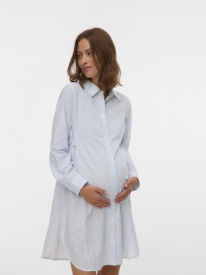 Gravid kvinde i en løs, lyseblå, stribet kjole med knapper, der blidt holder sin mave og kigger lidt til siden.