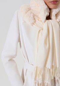 Manteau blanc avec un col en peluche beige clair. Une écharpe crème drape autour du cou, présentant des bords frangés et une texture douce et lisse.