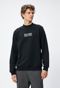Czarna bluza z ribbowanym dekoltem typu crew, z białym napisem "NEW YORK" na przodzie w wyrazistym kroju. Gładka tekstura i dopasowane mankiety przy nadgarstkach oraz u dołu.