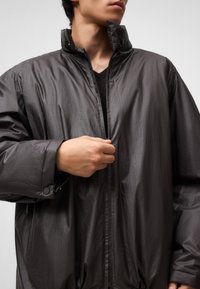 Oversized kurtka w kolorze węgorzowym, wykonana z lekkiego, błyszczącego materiału, z wysokim kołnierzem i zapięciem na zamek błyskawiczny z przodu. Posiada mankiety zapinane na napy.