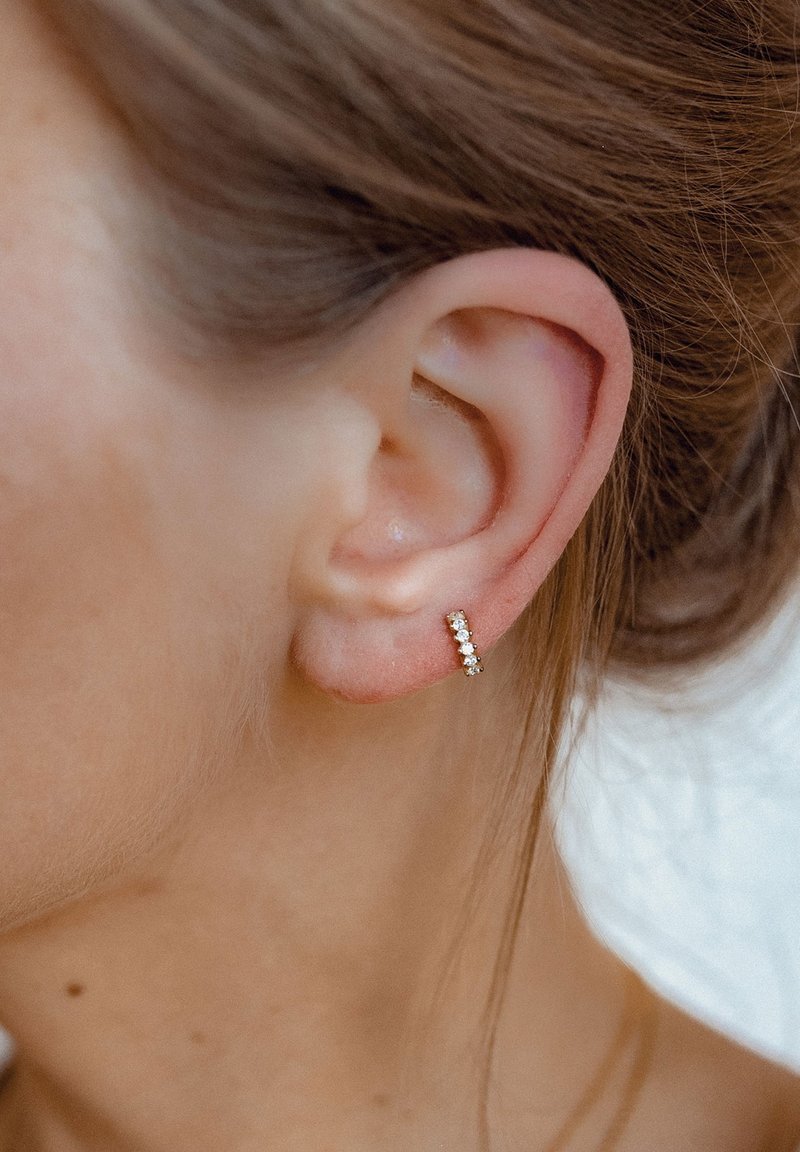 Boucle d'oreille en or avec cinq petites pierres précieuses transparentes disposées en ligne, posée contre l'oreille d'une personne ayant les cheveux châtains clairs.