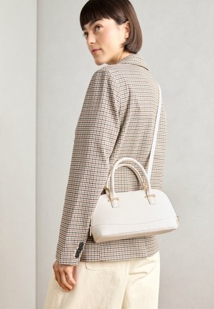 Woman wearing a checkered blazer and light pants carries a small white handbag with handles and a shoulder strap on her back.