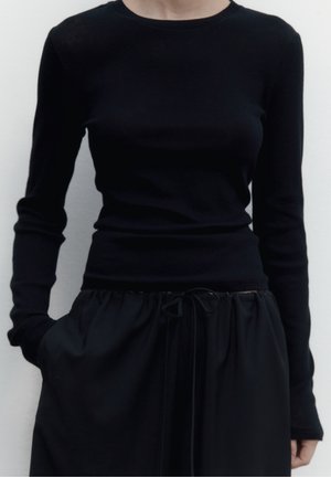 Femme portant un top noir moulant à manches longues, rentré dans un pantalon large noir avec un cordon de serrage, debout avec une main dans la poche.