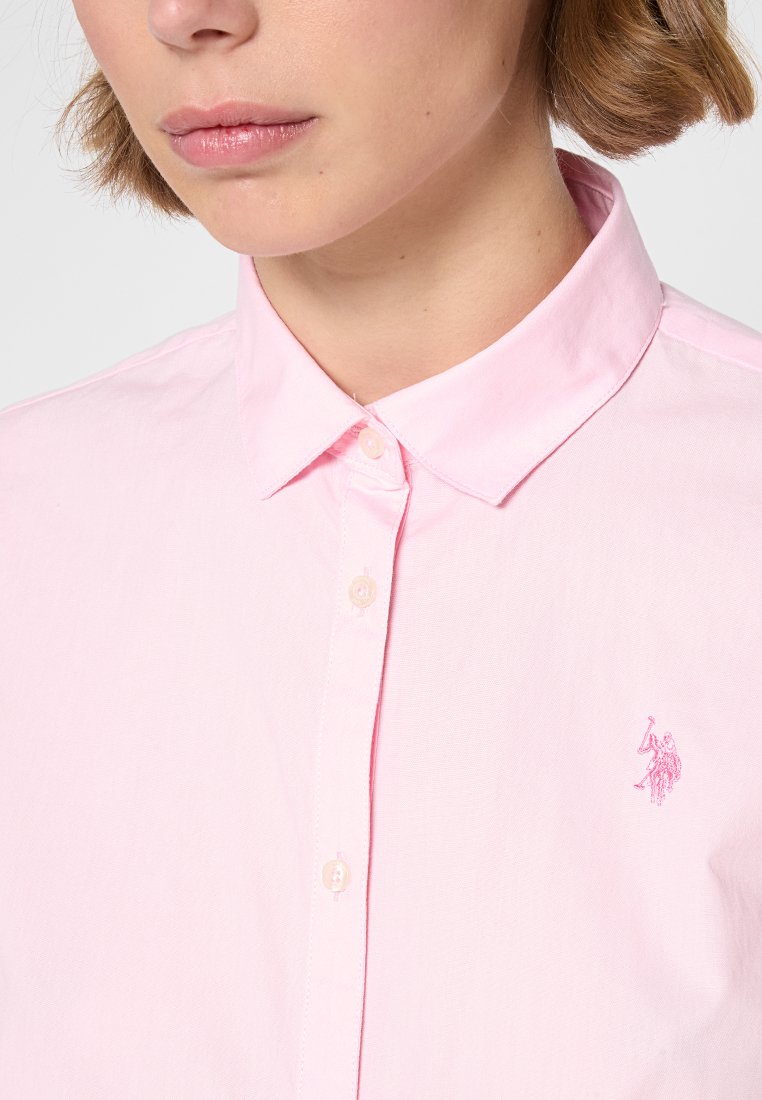 Chemise boutonnée rose clair avec col, en coton. Présente un petit logo brodé sur la poitrine gauche. Texture lisse.