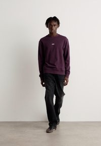 Sudadera púrpura con cuello redondo y un pequeño logo, combinada con jeans oscuros y zapatos, modelo caminando frente a un fondo liso.