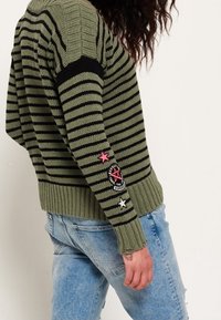 Maglione a righe verde oliva e nero con orlo a coste, caratterizzato da toppe a forma di stella rosa e bianca e un logo circolare sulla manica.