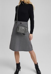 Šedá sukně v herringbone vzoru s černými knoflíky, spárovaná s černým rolákem. Doplňky zahrnují šedou crossbody kabelku s texturou a černé boty.