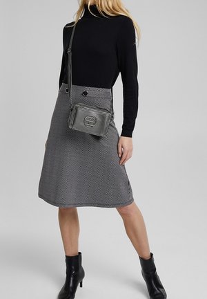 Šedá sukně v herringbone vzoru s černými knoflíky, spárovaná s černým rolákem. Doplňky zahrnují šedou crossbody kabelku s texturou a černé boty.