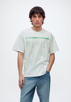 Ung mand med mørkt bølget hår bærer en hvid T-shirt med grøn japansk tekst og falmede linjer, kombineret med blå denimjeans.