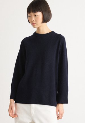 Jonge vrouw met kort donker haar, gekleed in een loszittende donkerblauwe trui en witte pants, tegen een effen achtergrond.