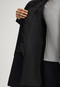 Cappotto nero con un'esterno liscio e un rivestimento imbottito nero, dotato di una cerniera e un'apertura frontale, che rivela uno strato interno grigio.