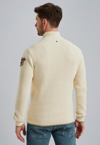 Crème ribbeltrui met hoge kraag, voorzien van een textuurafwerking en een badge op de linker mouw. Gedragen met blauwe jeans.