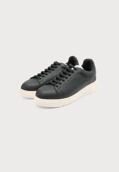 Paire de baskets en cuir noir avec semelles blanches épaisses et lacets noirs, placées côte à côte sur un fond clair uni.