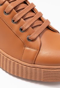 Gros plan sur une sneaker en cuir beige avec des lacets marron épais, des œillets en métal et une semelle en caoutchouc texturé avec des détails cousus.