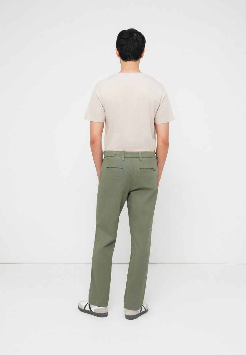 Hombre de espaldas vistiendo una camisa beige de manga corta, pantalones verde oliva con bolsillos traseros con cremallera y botones, y sandalias blancas.