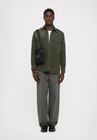 Olive green button-up shirt with two chest pockets, paired with gray wide-leg pants and black shoes. Includes a small black crossbody bag.